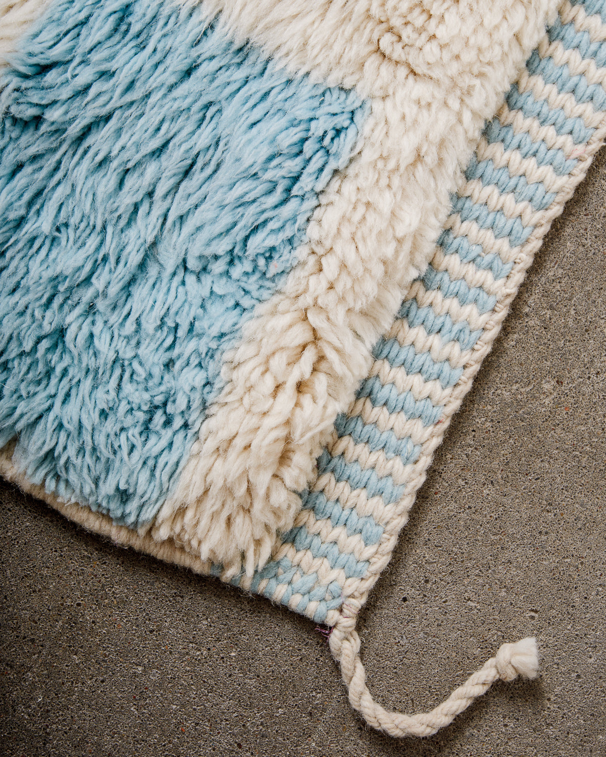 Corner of rug made of thick wool pile in sky blue and cream and finished with tightly woven end with alternating lines of blue and cream, and a single wool tassel on the corner.